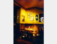 Ein gemütliches Kneipeninterieur mit Holztischen und -stühlen in einem warm beleuchteten Raum. An der gelblichen Wand hängen mehrere Bilder und ein rundes Schild mit der Aufschrift "Jever", daneben befindet sich eine kleine Tafel mit handgeschriebener Speisekarte.