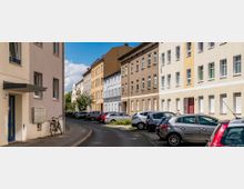Wohnstraße mit dicht parkenden Autos entlang beider Straßenseiten, umgeben von mehrstöckigen Wohnhäusern in hellen Farben. Einige Gebäude haben geöffnete oder geschlossene Fensterläden, und ein Fahrrad lehnt an der Fassade eines Hauses links im Bild.