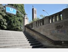 Steintreppe mit Geländer führt zur U-Bahn-Station „Hohenzollernplatz“, erkennbar durch ein hängendes blaues Schild. Im Hintergrund sind Bäume und der Turm einer Kirche mit Kreuz und Uhr zu sehen.