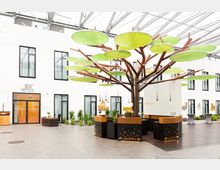 Moderner Hotel-Innenbereich mit Glasdach, einer Rezeption in Baumform mit braunem Stamm und großen grünen, blattförmigen Elementen darüber. Im Hintergrund weiße Wände mit symmetrischen Fenstern und einer gläsernen Eingangstür zur "Moa Bar" und "Convention Hall".