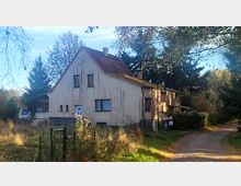 Ein schlichtes, zweistöckiges Wohnhaus mit Putzfassade und rotem Satteldach, umgeben von Bäumen und einem Garten. Im Vordergrund verläuft ein gepflasterter Weg, und ein Zaun grenzt das Grundstück ab.