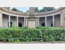 Halbkreisförmige Steinarchitektur mit Säulen und verzierten Bögen, davor eine begrünte Hecke und ein steinernes Denkmal in der Mitte; im Hintergrund sind Bäume zu sehen.