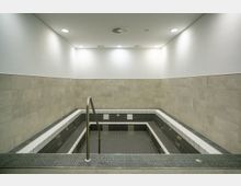 Innenraum eines modernen Tauchbeckens mit grauen Fliesen an den Wänden und dunklen Fliesen im Becken. An der Vorderseite führt ein Metallhandlauf ins rechteckige Becken, umgeben von weißer und schwarzer Kante.