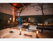 Gemütlicher, rustikaler Innenraum mit hohen Decken, Holzbalken und einem dekorierten Essbereich. Die Tische sind mit Kerzen und Speisen gedeckt, umgeben von Pflanzen und Wanddekorationen auf grünen Wänden.