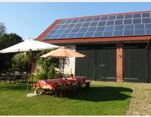 Ein rustikaler Hof mit einer Scheune aus Backstein und Holz, auf deren Dach Solarpaneele installiert sind. Im Vordergrund stehen Gartenmöbel und zwei Sonnenschirme auf einer Rasenfläche.