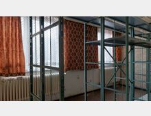 Ein leerer Raum mit hohen Fenstern, die von orange-gemusterten Vorhängen und weißen Gardinen bedeckt sind. Metallregale sind im Vordergrund, während unter den Fenstern weiße Heizkörper erkennbar sind.