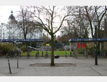 Gepflasterter Platz mit einem Baum in der Mitte, umgeben von Fahrradständern unter überdachten Unterständen. Im Hintergrund ein weißes historisches Gebäude und eine Baustelle hinter einem Metallzaun, daneben einige Parkflächen und Büsche.
