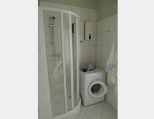 Badezimmer mit einer weißen, halbkreisförmigen Duschkabine mit Glaswänden auf der linken Seite und einer weißen Waschmaschine mit einem kleinen Boiler darüber auf der rechten Seite. Die Wände und der Boden sind mit hellen Fliesen versehen, und ein Handtuch hängt neben der Waschmaschine.