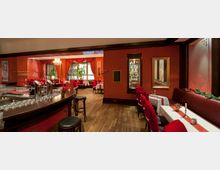 Ein elegant eingerichtetes Restaurant mit rotem Wandanstrich, roten Polsterstühlen und weißen Tischdecken. Im Vordergrund befindet sich eine Bar mit Hockern, im Hintergrund sind festlich dekorierte Tische, große Fenster und rote Vorhänge sichtbar.