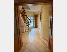 Eingangsbereich eines Hauses mit hell gefliesten Böden, einer Holztreppe mit schmiedeeisernem Geländer, einer Tür mit Glaseinsätzen und einer weiteren Holztür. Die Wände sind beige und leicht strukturiert.