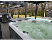 Outdoor patio with a large white hot tub on a dark deck beneath a modern metal pergola, with a dining table and chairs nearby. In the background are a lawn bordered by tall shrubs and stone privacy walls, with a wooded area beyond.