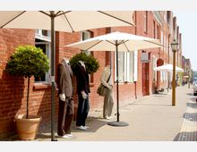 Straßenszene mit roten Backsteingebäuden, vor denen Schaufensterpuppen in Anzügen und Freizeitkleidung sowie Topfpflanzen unter großen Sonnenschirmen stehen. Im Hintergrund weitere Geschäfte mit offenen Fenstern und Gehweg.