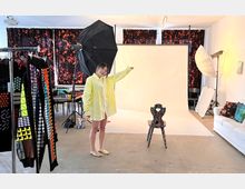 Ein Fotostudio mit einer hellen Leinwand als Hintergrund, umgeben von Beleuchtungsequipment wie Softboxen und Studiolampen. Im Vordergrund stehen ein gemusterter Stuhl und bunte Kleidungsstücke auf einem Kleiderständer, während eine Person in einem gelben Hemd den Bereich präsentiert.