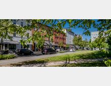 Stadtstraße mit mehrstöckigen Wohnhäusern in verschiedenen Farben und Geschäften im Erdgeschoss, flankiert von Bäumen. Im Vordergrund ist eine Grünfläche mit Rasen und Bäumen, während Autos und Fußgänger die Straße beleben.