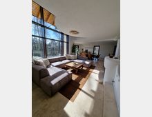 Wohnzimmer mit großen bodentiefen Fenstern, die viel Tageslicht hereinlassen, einer großen grauen Eckcouch auf einem dunklen Holzboden, einem niedrigen Holztisch und einem Esstisch mit Stühlen im Hintergrund. Die Decke ist mit geschwungenen Formen gestaltet, und der Boden ist teilweise mit hellen Fliesen ausgelegt.