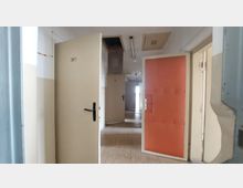 Korridor mit mehreren geöffneten Zimmertüren, nummeriert unter anderem mit "361" und "360", und Wänden in cremefarbenem Anstrich. Im Hintergrund ist eine schwere, orangefarbene Tür und ein heller Lichtschacht sichtbar; die Decke zeigt Lüftungsgitter und Leuchtstofflampen.