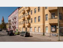 Straßenecke mit gelbem Wohngebäude und rosa-weißem Altbau im Hintergrund, beide mit mehreren Balkonen. Im Vordergrund parken Autos entlang der Straße, während links ein Kirchturm und Bäume sichtbar sind.