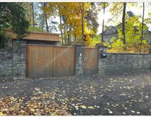 Steinerne Grundstücksmauer mit einem großen Holztor und einer kleineren Tür, beide mit geometrischen Mustern verziert; im Hintergrund sind herbstliche Bäume und ein Wohnhaus zu sehen.