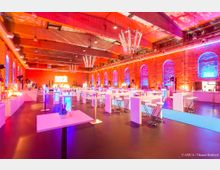 Großer Veranstaltungsraum mit Backsteinwänden, hoher Holzdecke und modernen Leuchtern. Die Halle ist mit hohen Tischen und Barhockern eingerichtet, in einem farbigen Lichtmix aus Blau, Rot und Orange beleuchtet.