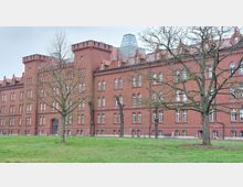 Historisches rotes Backsteingebäude mit zahlreichen schmalen, bogenförmigen Fenstern und einem modernen gläsernen Dachaufsatz. Im Vordergrund befindet sich eine Grünfläche mit einigen kahlen Bäumen.