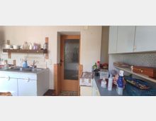 Kleine, schlichte Küche mit weißen Schränken und einem Doppelspülbecken. An der Wand befindet sich ein Holzregal, daneben eine Tür mit Milchglasscheibe; auf den Arbeitsflächen stehen Reinigungs- und Alltagsgegenstände.