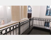 Obergeschoss eines Gebäudes mit cremefarbenen Rundsäulen, einem schwarzen Metallgeländer mit kreisförmigen Verzierungen und großen Fenstern, die auf eine städtische Umgebung blicken. Die Wände sind weiß, mit minimalistischen architektonischen Details.