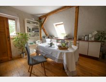 Gemütlicher Küchen- und Essbereich im Dachgeschoss mit sichtbaren Holzbalken, Holzfußboden und weiß verputzten Wänden. Ein runder Tisch mit Tischdecke steht vor einer Küchenzeile unter einem Dachfenster; daneben ein offenes Regal, Zimmerpflanzen und ein seitliches Fenster.
