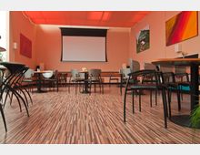 Ein moderner Veranstaltungsraum mit holzgemustertem Laminatboden, runden Tischen und verschiedenen Stuhlmodellen. An der Vorderwand befindet sich eine weiße Projektionsleinwand, die von bunten Bildern umgeben ist, während die Decke mit roten Segeltuchbahnen bespannt ist.