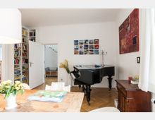 Wohnzimmer mit heller Einrichtung, einem großen schwarzen Flügel und Holzmöbeln. An der Wand befinden sich ein Regal mit Büchern und Spielen, eine Bildercollage sowie ein rotes Gemälde; eine offene Tür führt zu einer Treppe im Hintergrund.