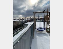 Schneebedeckte Dachterrasse mit Blick über eine Stadt, im Hintergrund sind die Skyline mit Fernsehturm und weiteren hohen Gebäuden sichtbar. Im Vordergrund steht ein Holzgerüst mit einer blauen Nestschaukel.