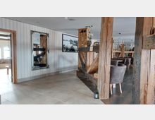 Innenraum eines Restaurants mit rustikalem Design, geprägt durch Holzpfeiler, Holzbalken und eine Kombination aus moderner und traditioneller Dekoration. Zu den Details gehören ein großer Spiegel an einer weiß gestreiften Wand, eine Schwarz-Weiß-Fotografie, Kronleuchter und gepolsterte Sessel um Esstische.