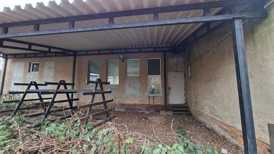 Überdachter Innenhof oder Anbau mit Metallträgern und Wellblechdach vor einer verputzten Gebäudewand mit mehreren Fenstern und einer Tür. Der Boden ist erdig und von Gestrüpp sowie Unrat bedeckt, daneben stehen schwarze Metallrahmen oder Gestelle.