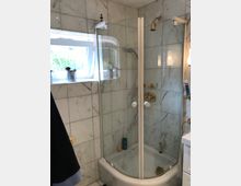 Duschkabine mit gebogenen Glastüren in einem Badezimmer mit weißen Marmorkacheln. Links ein kleines, geöffnetes Fenster, rechts ein Waschbecken mit Wandbeleuchtung.