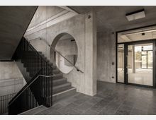 Minimalistischer Treppenhausbereich aus Beton mit schwarzem Metallgeländer, einem kreisförmigen Wanddurchbruch und großen Glastüren, die zu einem hellen Raum führen. Der Boden besteht aus dunklen Steinfliesen.