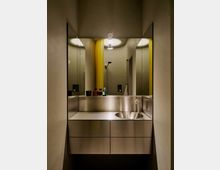 Schmales, modernes Badezimmer mit Edelstahl-Waschtisch und rundem eingelassenem Becken, flankiert von großen Spiegeln an beiden Seiten. Im Hintergrund sind eine Dusche, gelbe Vorhänge und runde Deckenöffnungen mit Beleuchtung zu sehen.