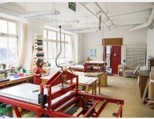 Ein heller Arbeitsraum mit großen Fenstern, in dem Druckmaschinen und Werkbänke stehen. Eine Person arbeitet an einem der Tische, umgeben von Werkzeugen und Materialien; im Hintergrund sind rote Türen und Regale sichtbar.