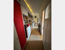 Ein schmaler Hausflur mit Hochregalen an der linken Wand, die Bücher und Dekorationsgegenstände enthalten. Rechts befindet sich eine weiße Tür und ein Spiegel an der Wand, während am Ende des Flurs ein Hund steht. Der Flur ist mit Teppich ausgelegt und von warmer Beleuchtung erhellt.