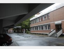 Zweistöckiges Gebäude aus rotem Backstein mit großen Fenstern, angeschrieben als „Haus der Kirche“. Im Vordergrund überdachter Gehweg, nasser Asphalt und ein geparkter roter Wagen unter Betonträgern, Hintergrund mit Bäumen und weiteren Gebäuden.