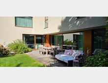 Moderner Gartenbereich mit Holzterrasse, großen Schiebetüren aus Glas und einer beige verputzten Hausfassade. Die Terrasse ist mit Esstischen, Stühlen und einer bequemen Sitzgelegenheit ausgestattet, umgeben von Pflanzen und einer Rasenfläche.