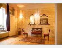 Ein rustikaler Raum mit abgeblätterten Wänden, einer hölzernen Sitzgruppe mit roter Polsterung, drei antiken Wandspiegeln und zwei Teppichen auf Holzboden. Links befinden sich ein Fenster mit roten Vorhängen, ein Heizkörper und Pflanzen auf einem Regal.