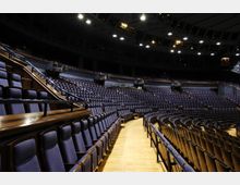 Großes Theater- oder Konzertsaal-Interieur mit mehreren Sitzreihen in einem halbkreisförmigen Design, blauen Polstersitzen und Holzböden. Die Decke ist mit Lichtern und akustischen Elementen ausgestattet.
