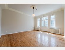 Heller, leerer Altbau-Raum mit breiten Holzdielen, hohen Decken mit Stuck und einer zentralen Pendelleuchte. An der Stirnseite befinden sich hohe, weiß gerahmte Fenster und eine verglaste Flügeltür, darunter weiße Heizkörper.