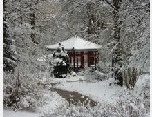 Schneebedeckter Garten mit einem roten, asiatischen Pavillon in der Mitte, umgeben von verschneiten Bäumen und Sträuchern. Ein gewundener Fußweg führt durch die winterliche Landschaft.