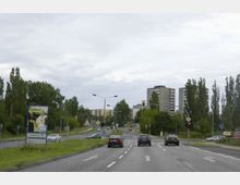 Straßenkreuzung mit mehreren Fahrspuren in einer städtischen Umgebung, umgeben von Bäumen und Wohnhochhäusern im Hintergrund. Verkehrsampeln leuchten rot, während Autos auf der Straße fahren, und ein Werbeschild mit Willkommenstext steht am Straßenrand.