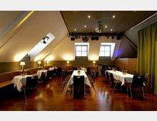 Gastraum unter einem schrägen Dach mit Dachfenstern, warmem Licht und Holzboden, ausgestattet mit Tischen mit weißen Tischdecken und schwarzen Stühlen. Entlang der Wände stehen gepolsterte Sitzbänke mit kleinen Tischlampen; an der Decke sind Spotlights und eine Discokugel angebracht.