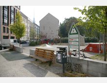 Öffentlicher Spielplatz mit Sitzbänken, Mülleimer und einer Hinweistafel, umgeben von Wohnhäusern und Bäumen; im Hintergrund ist ein modern gestaltetes Gebäude mit einer Wandzeichnung sowie der Berliner Fernsehturm zu sehen.