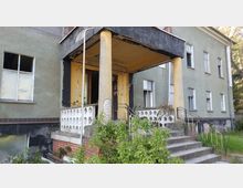 Eine verlassene Eingangsterrasse eines Gebäudes mit gelb gestrichenen Säulen und verblasster Fassade. Eine Treppe mit Geländer führt zur verfallenen, teilweise bewachsenen Terrasse, während Fenster mit teils beschädigtem Glas die grüne Außenwand des Gebäudes durchbrechen.