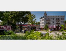 Straßenszene mit einem mehrstöckigen Gebäude mit heller Fassade und Ziegeldach auf der rechten Seite, einer mit Graffiti versehenen kleinen Hütte auf der linken Seite und einer Bahnbrücke im Hintergrund, auf der ein roter Regionalzug fährt. Im Vordergrund befinden sich Bäume, Büsche und eine Grünfläche.