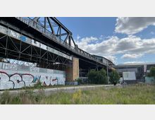 Industriebrücke mit Stahlträgern und Gerüstkonstruktionen, umgeben von Wänden mit Graffiti. Im Vordergrund befindet sich eine unbebaute Grünfläche, im Hintergrund sind moderne Gebäude und ein blauer Himmel mit Wolken sichtbar.