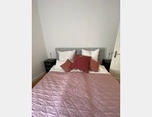 Ein Schlafzimmer mit einem Bett in der Mitte, das mit einem rosa gesteppelten Bettbezug bedeckt ist. Das Bett hat ein graues Kopfteil, weiße Kissen und dekorative, rot-rosa Kissen; daneben stehen zwei schwarze Nachttische mit Tischlampen.